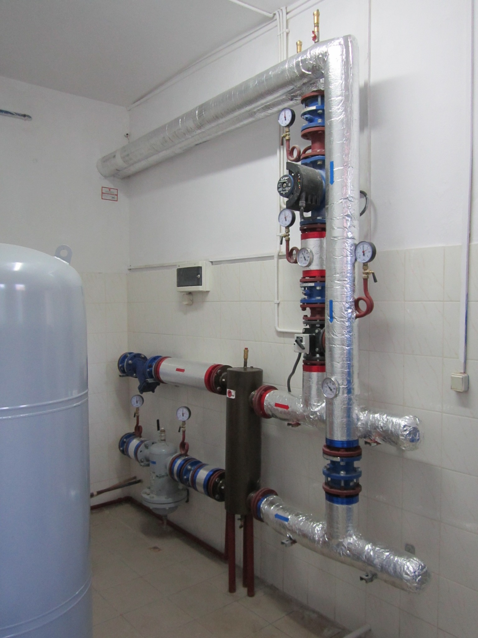 Złożona instalacja hydrauliczna z izolowanymi rurami, manometrami i pompą obiegową w pomieszczeniu z białymi płytkami.