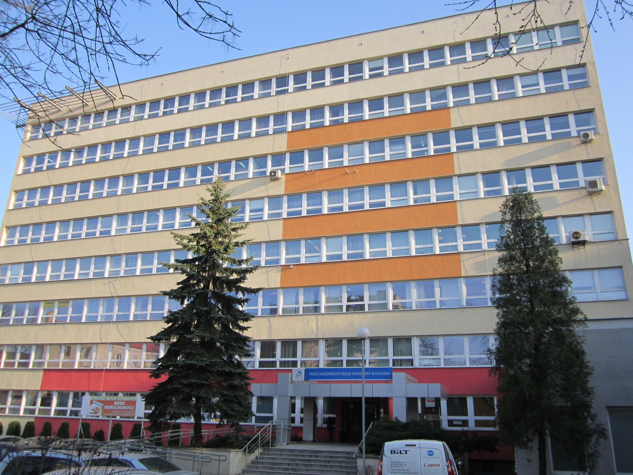 Wielopiętrowy budynek komercyjny z licznymi oknami, klimatyzatorami na fasadzie, dwoma drzewami iglastymi i zaparkowanym samochodem dostawczym z logo firmy BILT.