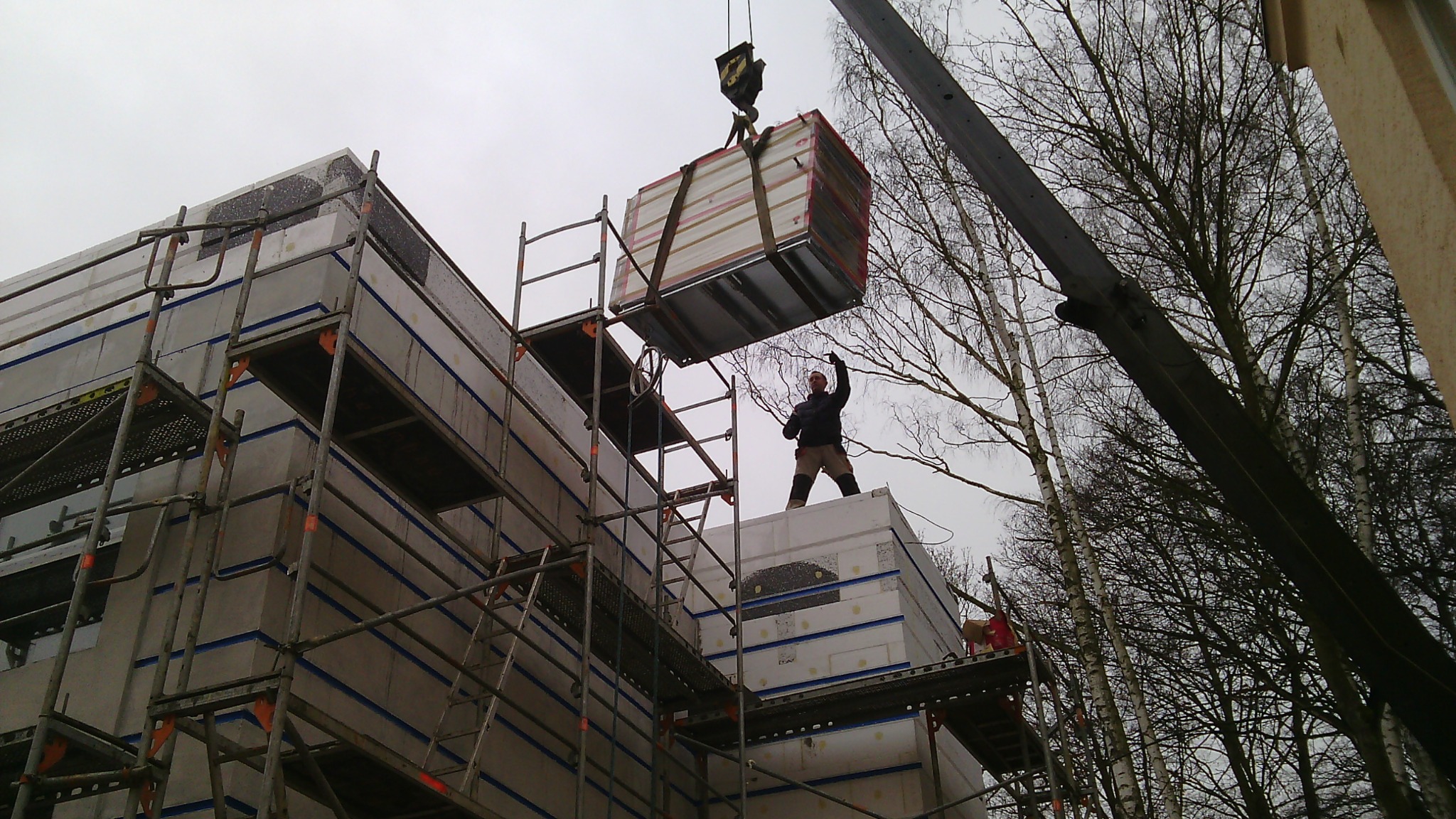 Montaż prefabrykowanych elementów budynku na placu budowy, z użyciem dźwigu i rusztowań, pracownik w kasku nadzoruje proces.