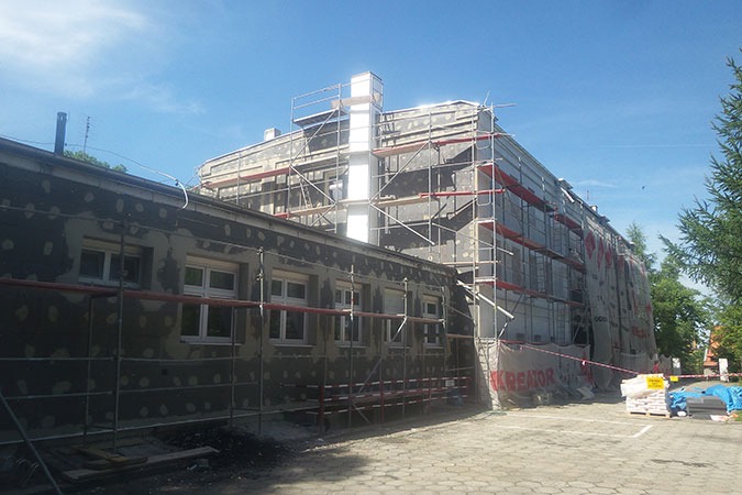 Budynek w trakcie ocieplania, widoczne rusztowania, elewacja z nałożonym klejem i siatką, częściowo zakryta siatką ochronną z logo, palety z materiałami budowlanymi.