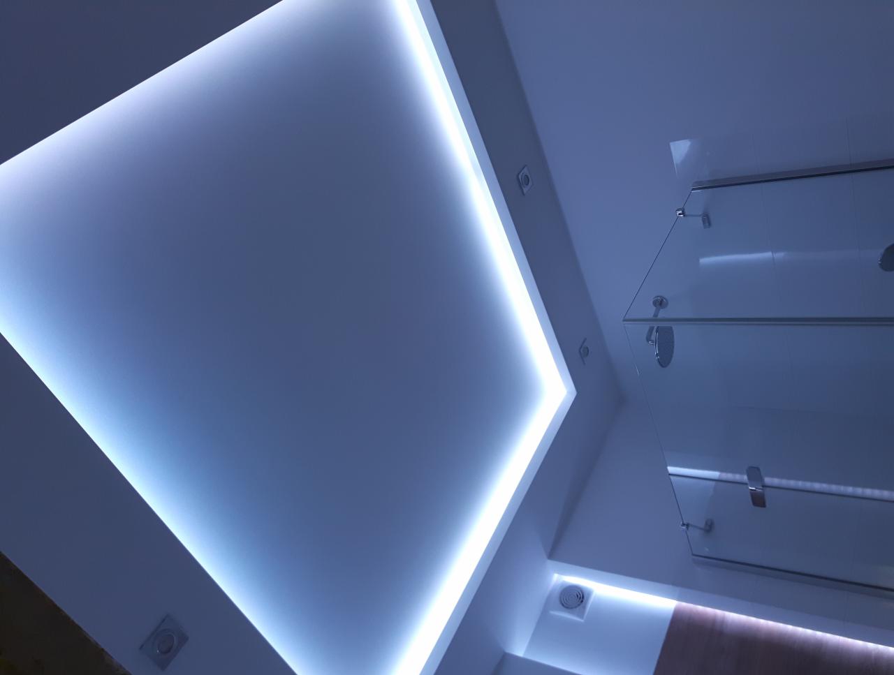 Nowoczesna łazienka z podświetlanym sufitem napinanym i szklaną kabiną prysznicową widziana z dołu.