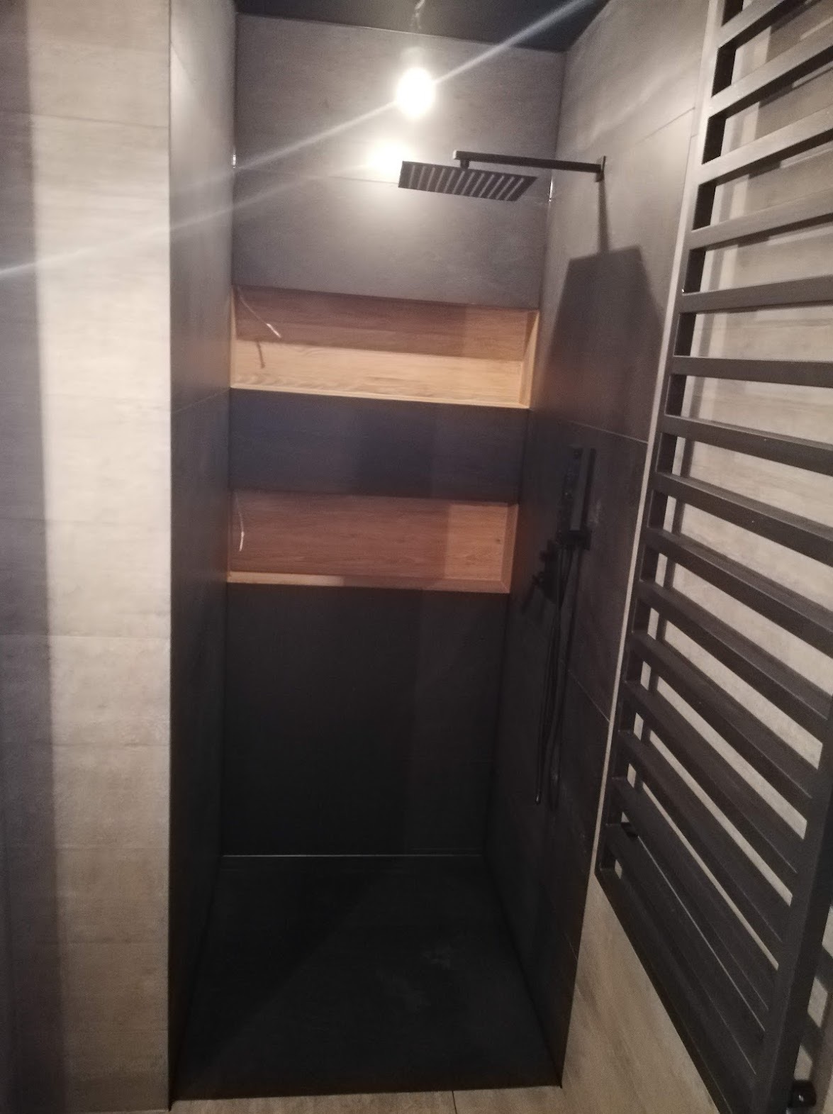 Nowoczesna kabina prysznicowa walk-in z ciemnymi płytkami, deszczownicą i drewnianymi półkami wnękowymi, widoczny grzejnik drabinkowy.