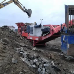 Żółta koparka CAT ładuje gruz do czerwono-białej kruszarki SANSVIK Q340, która przekazuje materiał do niebieskiej ciężarówki z kratą, na tle sterty ziemi i budynków.