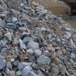 Kopalnia odkrywkowa: hałda szaro-brązowych kamieni o nieregularnych kształtach, z widocznym fragmentem czerwonego transportera taśmowego w tle, na zboczu ziemnym.