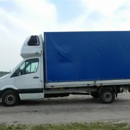 Biały bus z plandeką w intensywnym odcieniu niebieskiego, zaparkowany na szutrowej drodze, widok z boku, z podwyższoną kabiną sypialną.