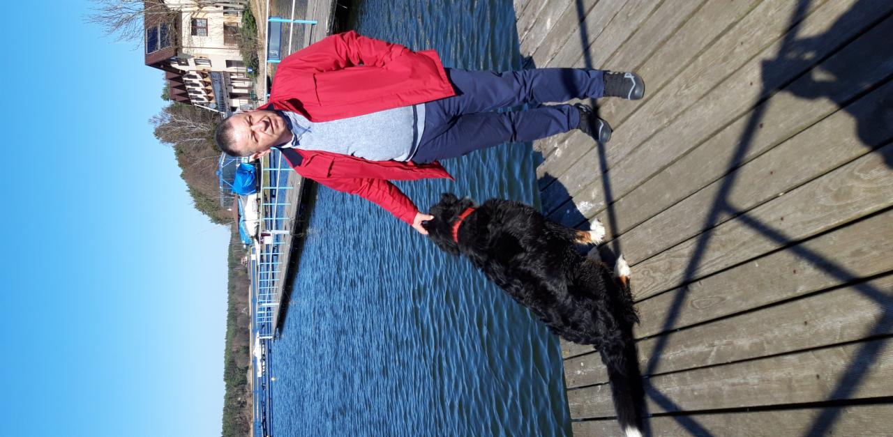 Mężczyzna w czerwonej kurtce głaszcze psa rasy berneński pies pasterski na drewnianym molo nad jeziorem w słoneczny dzień. W tle budynek i drzewa.