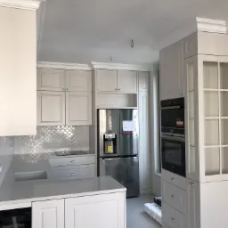 Jasna kuchnia z szarymi frontami, srebrną mozaiką nad blatem, zabudowaną lodówką i piekarnikiem oraz widocznymi elementami wykończeniowymi na podłodze, sugerującymi trwający montaż.