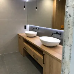 Nowoczesna łazienka z dwiema owalnymi umywalkami nablatowymi, drewnianym blatem z szafkami i szufladami, czarną armaturą oraz minimalistycznym oświetleniem wiszącym na tle szarej ściany z dużych...