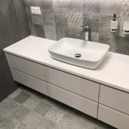 Nowoczesna łazienka z białą, prostokątną umywalką nablatową i szafką pod umywalką w kolorze białym, wzorzyste szaro-białe płytki na ścianie i podłodze, chromowana bateria i dozownik do mydła.