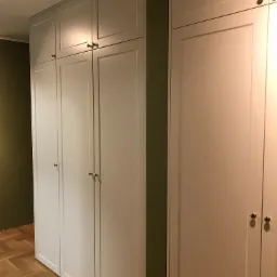 Wysoka, biała szafa wnękowa z złotymi gałkami, dopasowana do ściany w odcieniach oliwkowej zieleni, na parkiecie w jodełkę.