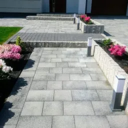 Szary chodnik z kostki brukowej prowadzący do domu z białymi ścianami, ozdobiony rabatami kwiatowymi i nowoczesnymi lampami.