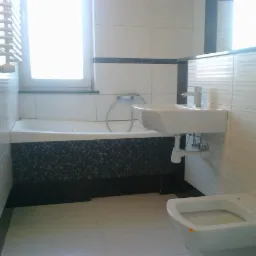 Wykończona łazienka z białą armaturą, wanną z mozaiką i umywalką. Ściany wyłożone białymi płytkami z szarym paskiem. Okno z żaluzjami w tle.