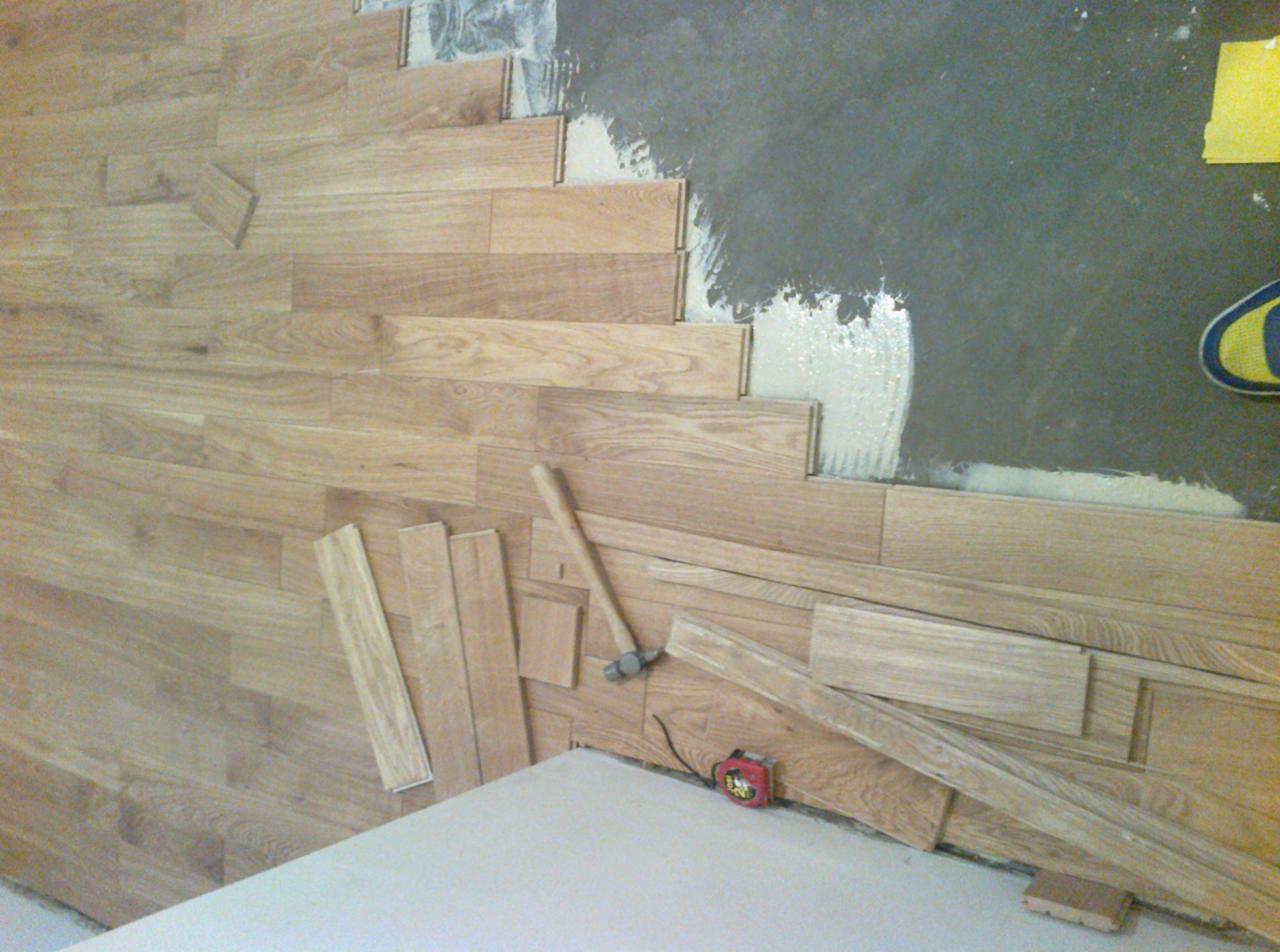 Układanie parkietu z jasnego drewna w trakcie prac. Widoczny klej, młotek i miarka. Częściowo ułożona podłoga i luźne deski. Ujęcie z góry, blisko podłoża.