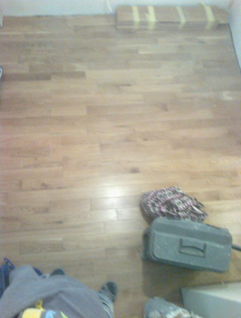 Jasne panele podłogowe po ułożeniu, widoczna skrzynka narzędziowa i częściowo osoba, na podłodze leżą panele zabezpieczone taśmą.