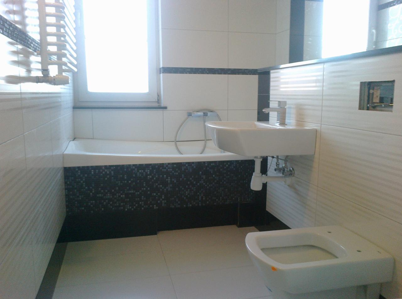 Wykończona łazienka z białą armaturą, wanną z mozaiką i umywalką. Ściany wyłożone białymi płytkami z szarym paskiem. Okno z żaluzjami w tle.