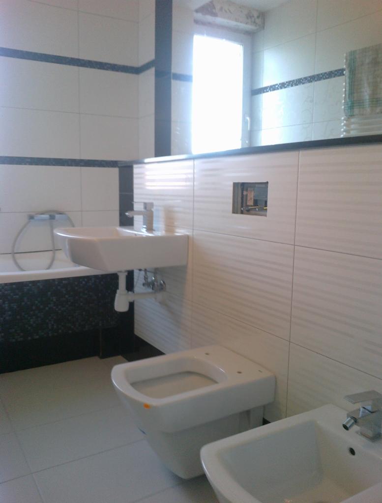 Nowoczesna, wykończona łazienka z białymi płytkami, armaturą i wiszącą toaletą. Minimalistyczny design, widoczne detale wykończeniowe i oświetlenie okienne.