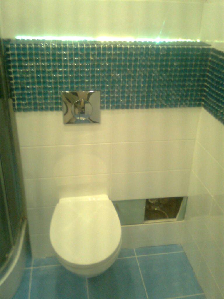 Biała toaleta podtynkowa z chromowanym przyciskiem spłukującym, wykończona mozaiką w odcieniach błękitu. Uzupełnieniem są jasne płytki na ścianie i podłodze.