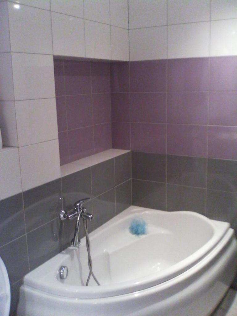 Narożna wanna z chromowaną armaturą na tle mozaiki z płytek w odcieniach bieli, szarości i fioletu. W tle wnęka wyłożona płytkami.