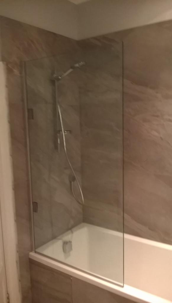 Nowoczesna kabina prysznicowa z pojedynczą szklaną ścianką, chromowaną armaturą i beżowymi płytkami imitującymi marmur. Widok na wannę i częściowo na baterię.