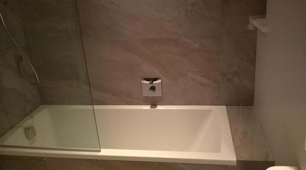 Biała wanna z szklanym parawanem i chromowaną armaturą na tle płytek imitujących marmur, tworząca elegancką strefę kąpielową w nowoczesnej łazience.