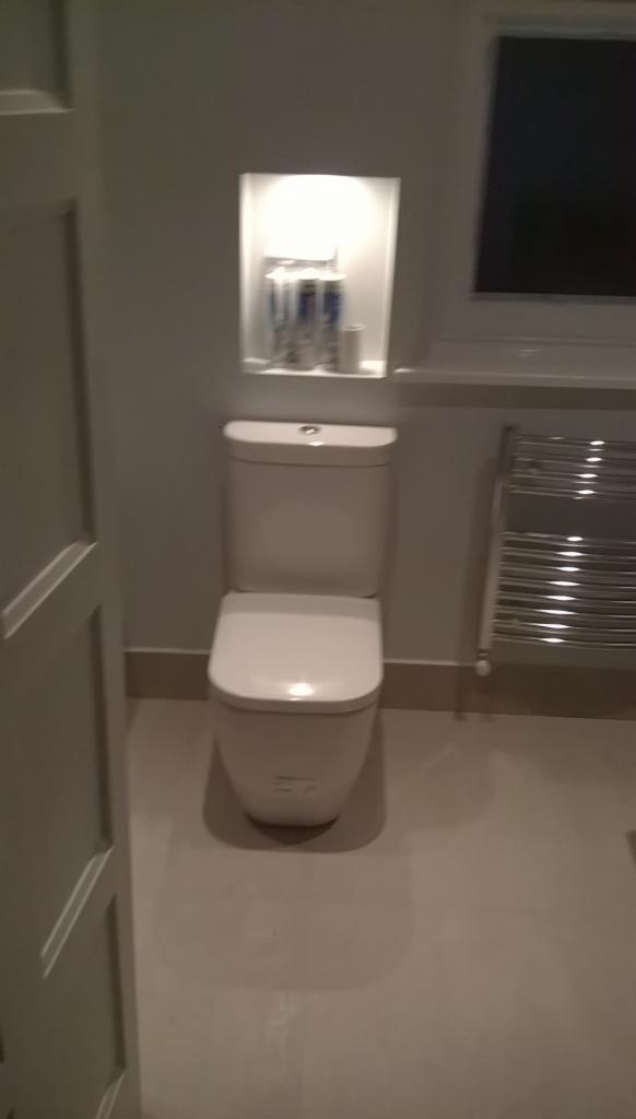 Biała toaleta z zabudowaną półką i grzejnikiem w wyremontowanej łazience. Widoczne okno i fragment drzwi. Całość utrzymana w jasnej kolorystyce.