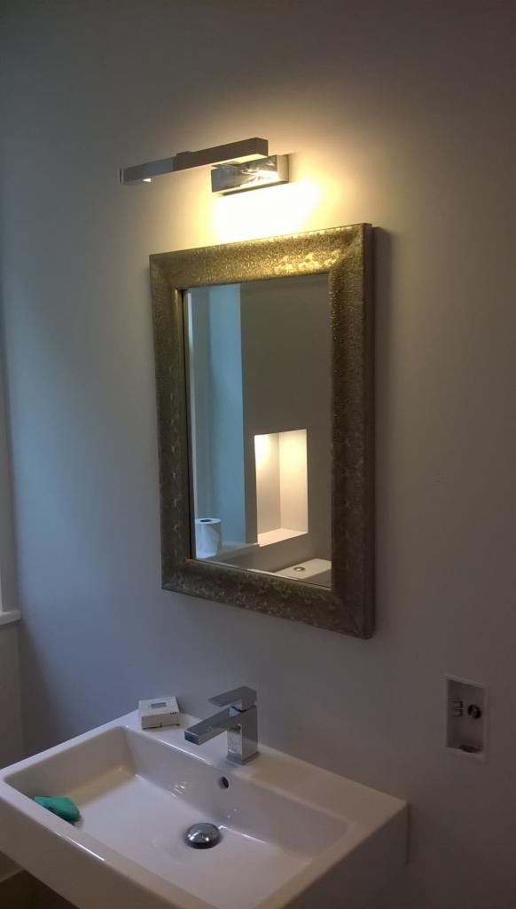 Umywalka z lustrem w złotej ramie i nowoczesnym oświetleniem. Widoczny fragment ściany z gniazdkiem elektrycznym. W lustrze odbicie toalety i wnęki.