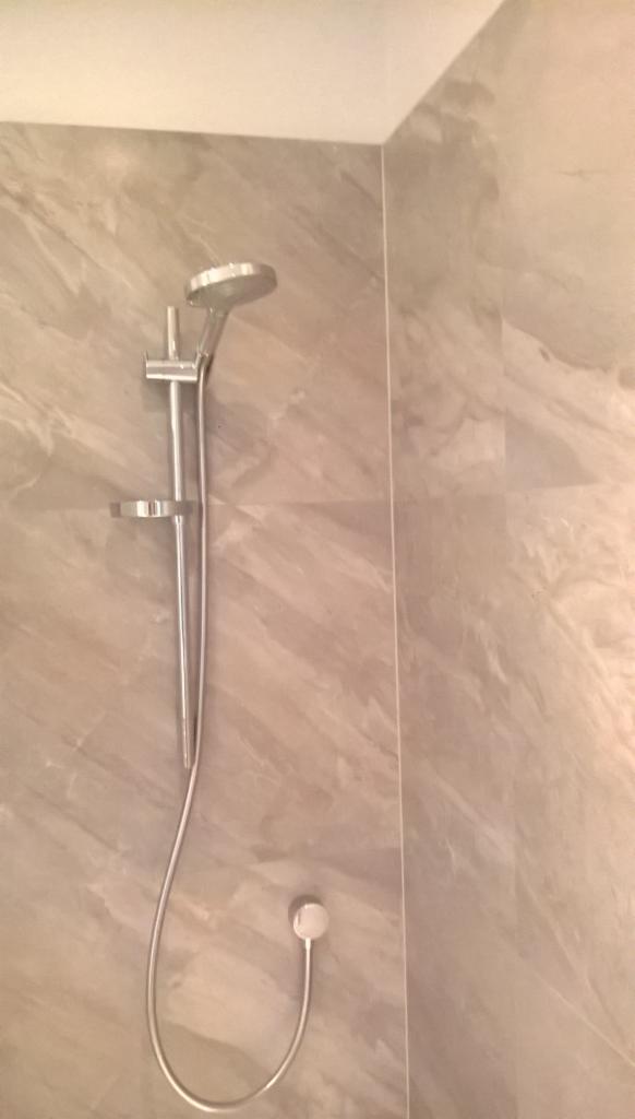 Błyszcząca, srebrna bateria prysznicowa zamontowana na ścianie z marmurkową, beżową płytką w wyremontowanej łazience. Widok z dołu, ukośny kadr.