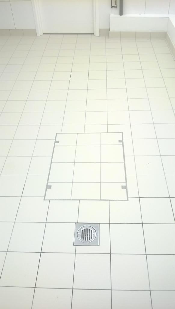 Biała, wykafelkowana podłoga z odpływem liniowym i ukrytą rewizją. Jasne, jednolite płytki z szarą fugą. Minimalistyczny design, czysta powierzchnia.