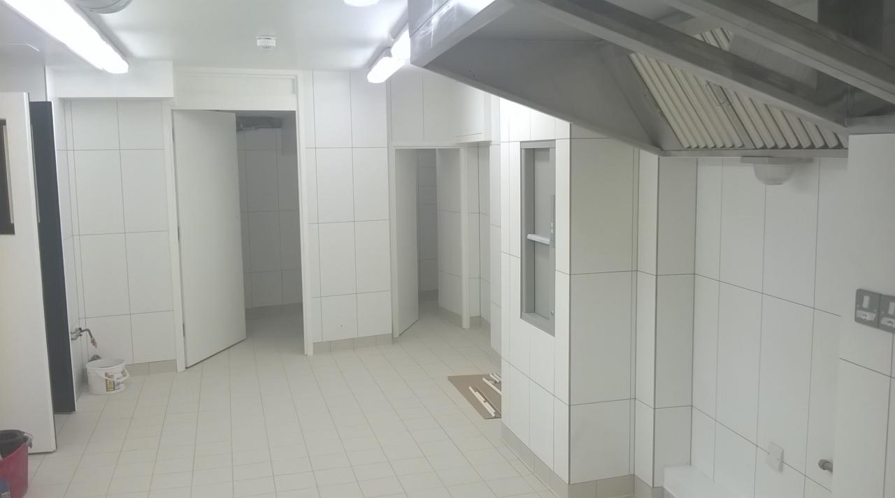 Biała, wykafelkowana kuchnia przemysłowa z otwartymi drzwiami i stalowym okapem. Widoczne gniazdka elektryczne i grzejnik. Czyste, jasne oświetlenie sufitowe.