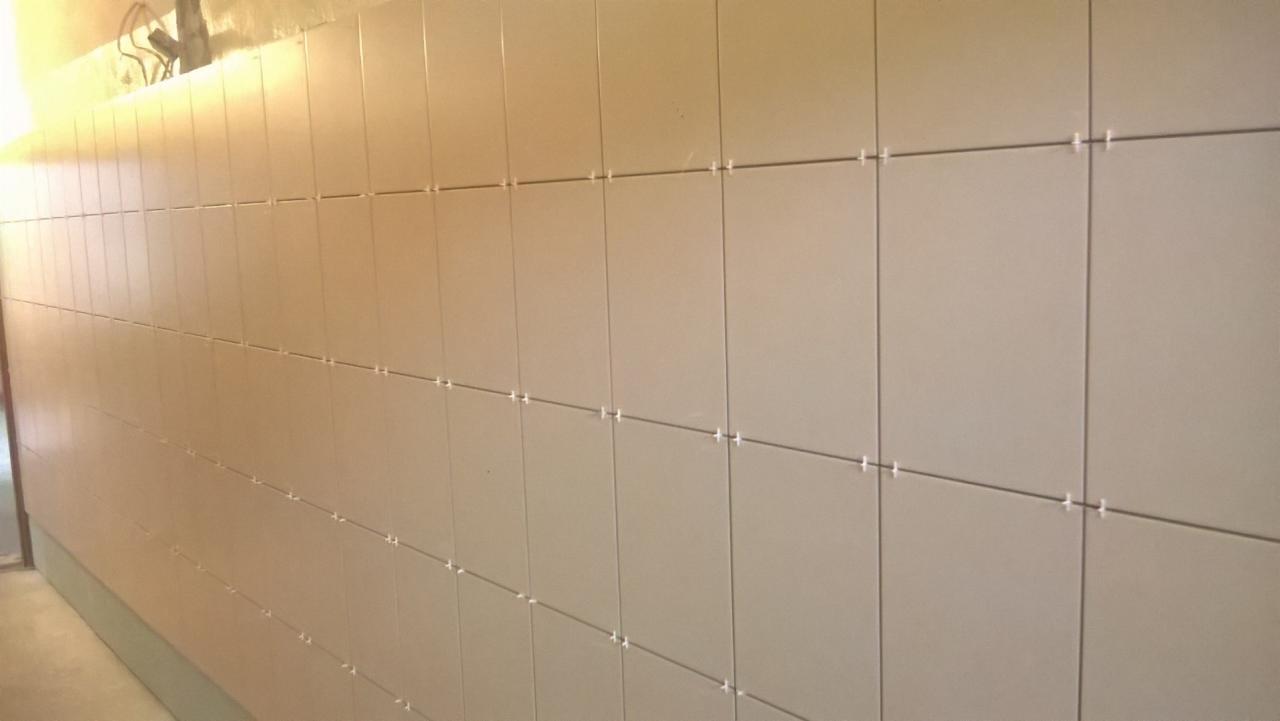 Ściana wyłożona beżowymi, kwadratowymi płytkami z plastikowymi krzyżykami dystansowymi, ukazująca etap prac wykończeniowych w pomieszczeniu.