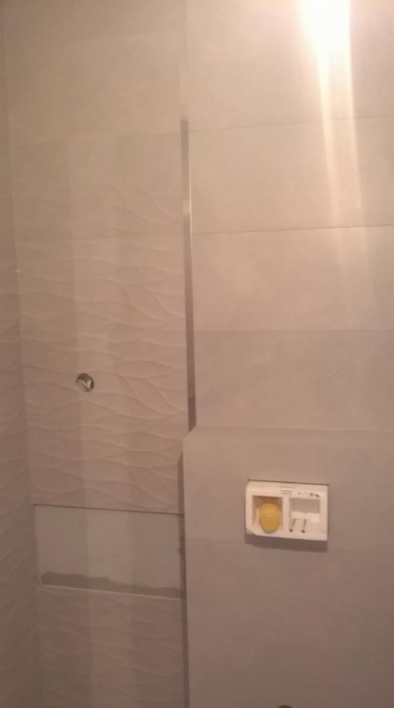 Wykończona łazienka z beżowymi płytkami o falistej strukturze. Widoczny przycisk spłuczki w zabudowie. Minimalistyczny design, nowoczesne wykończenie.