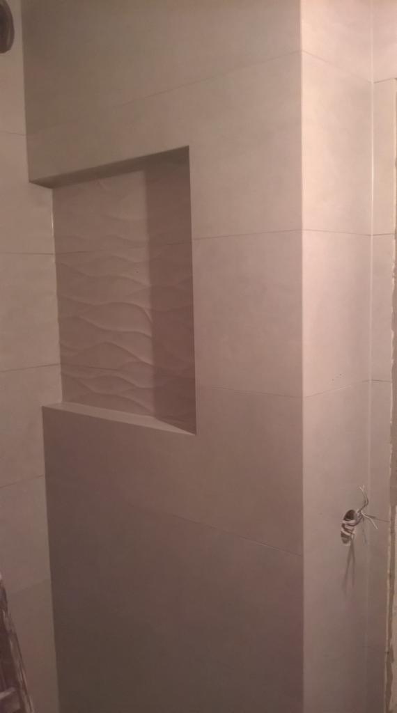 Wykończona wnęka z dekoracyjną płytką w łazience, obłożona jasnymi, beżowymi płytkami. Widoczne wystające przewody elektryczne.