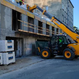 Żółta ładowarka teleskopowa JCB podnosi paletę białych bloczków silikatowych na budowę dwukondygnacyjnego budynku z szarego betonu komórkowego.
