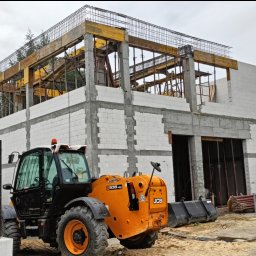 LEGO-BUD Piotr Legędź - Dwukondygnacyjny budynek w trakcie budowy z białych bloczków, z widocznym szalunkiem i zbrojeniami. Na pierwszym planie pomarańczowa ładowarka teleskopowa JCB.