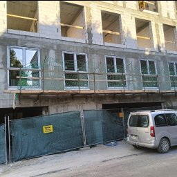 LEGO-BUD Piotr Legędź - Budynek w trakcie budowy z oknami i rusztowaniem, zabezpieczony siatką i ogrodzeniem. Widoczny samochód osobowy. Na górze budynku pracuje osoba.