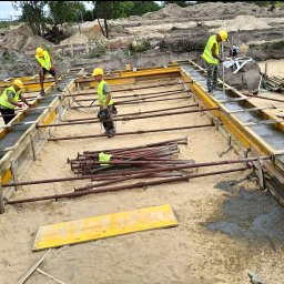 LEGO-BUD Piotr Legędź - Wylewanie fundamentów: czterech pracowników w żółtych kaskach i kamizelkach wylewa beton w szalunek. Na piasku leżą podpory i deski.