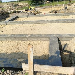 LEGO-BUD Piotr Legędź - Zalane betonem fundamenty domu, widok z góry na piasek wewnątrz. W tle ekipa budowlana i sprzęt na placu budowy. Kąt ujęcia podkreśla głębię wykopu.