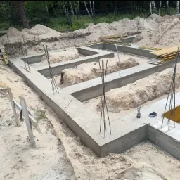 Zalane betonem fundamenty domu z wystającymi prętami zbrojeniowymi, widoczne drewniane szalunki i narzędzia. Tło to piasek i drzewa.