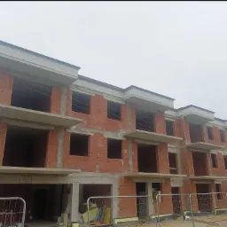 Wznoszenie trzypiętrowego budynku z czerwonej cegły, widoczne otwory okienne i balkony, zabezpieczone ogrodzeniem tymczasowym. Stan surowy otwarty.