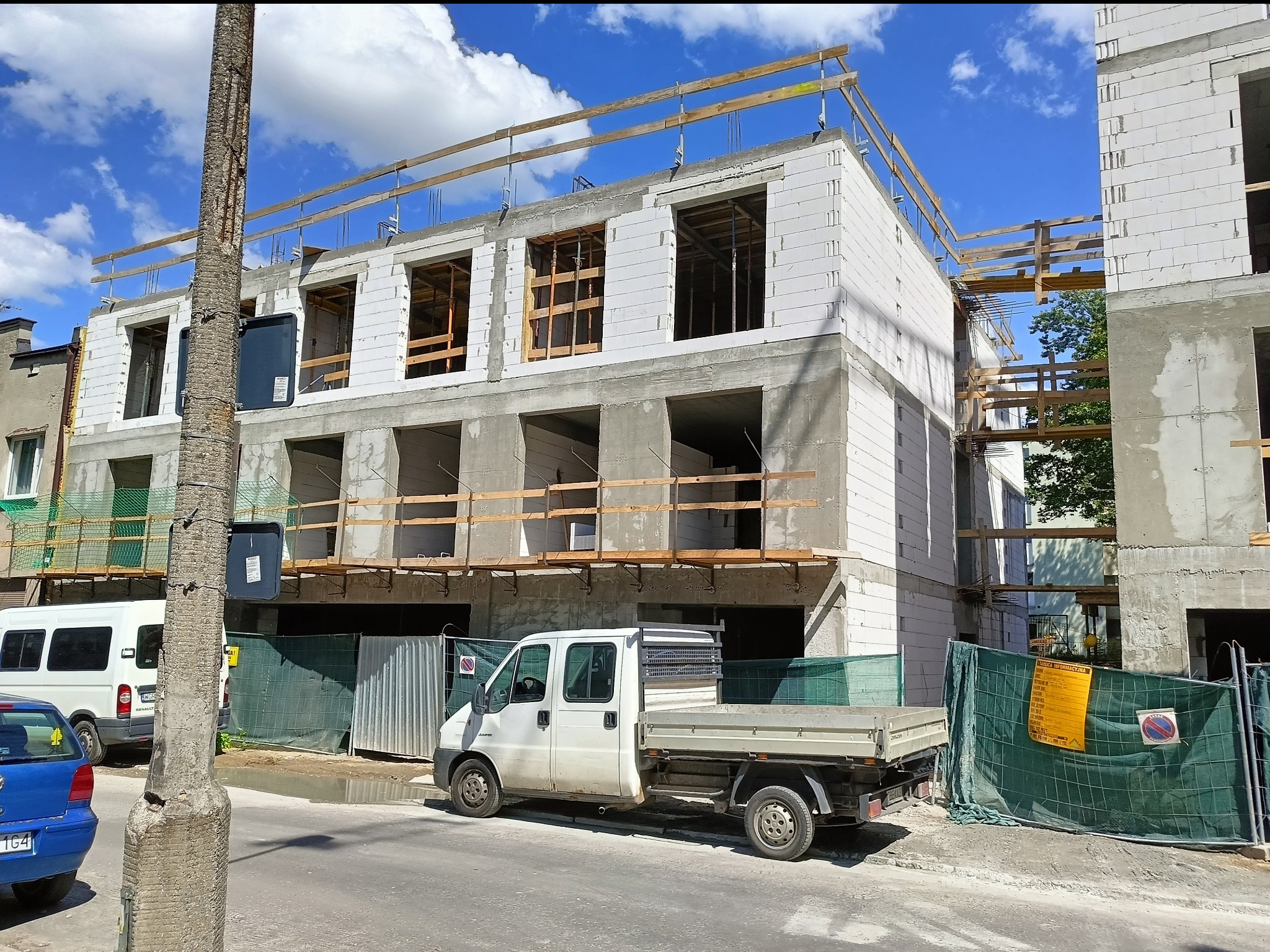 Budynek w stanie surowym otwartym z białego pustaka, widoczne drewniane stropy i zabezpieczenia. Na pierwszym planie zaparkowany biały samochód dostawczy.