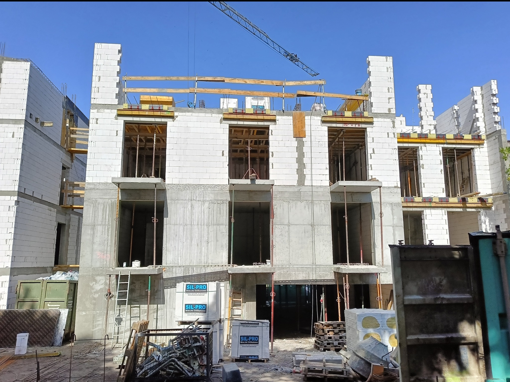 Budowa trzypiętrowego domu z białych bloczków silikatowych i betonowych elementów konstrukcyjnych. Widoczny żuraw budowlany na tle błękitnego nieba.