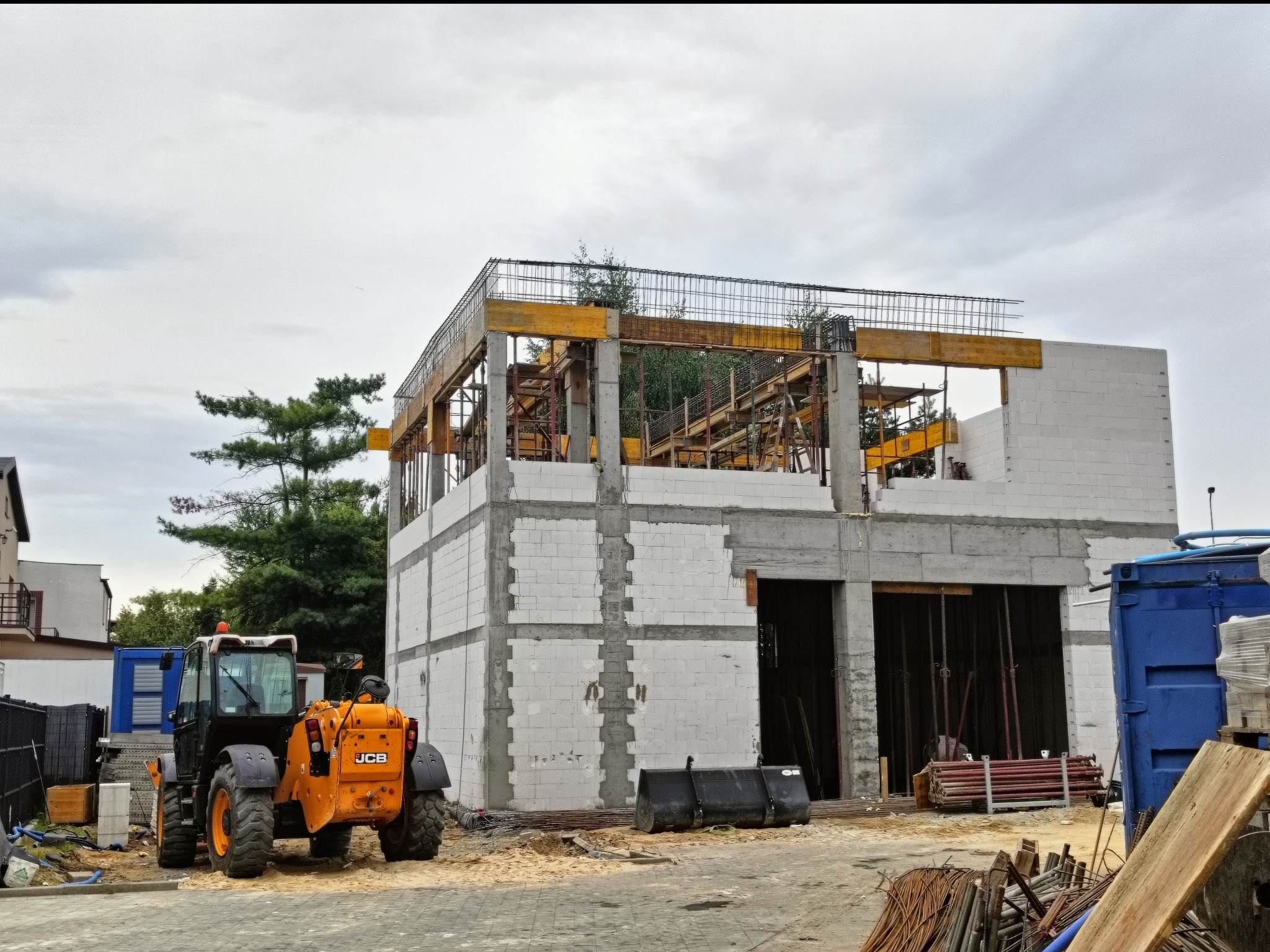 Budowa domu z białych bloczków, widoczny żuraw JCB i stalowe zbrojenia, betonowe filary. Ujęcie z zewnątrz, w tle zieleń drzew i fragmenty innych budynków.