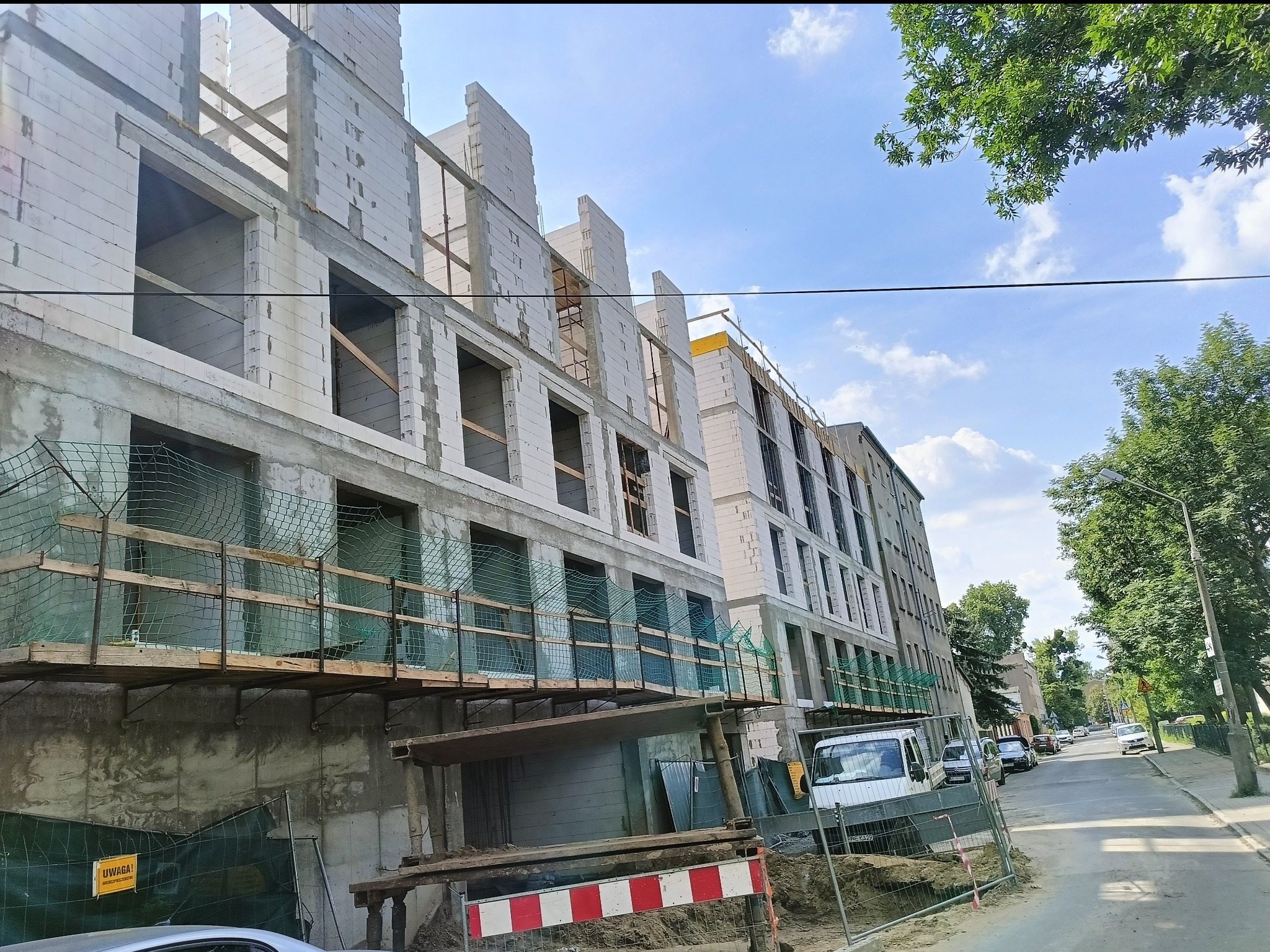 Wznoszenie wielopiętrowego budynku z białych bloczków, widok z dołu na elewację w trakcie prac murarskich, rusztowania zabezpieczone siatką, błękitne niebo w tle.