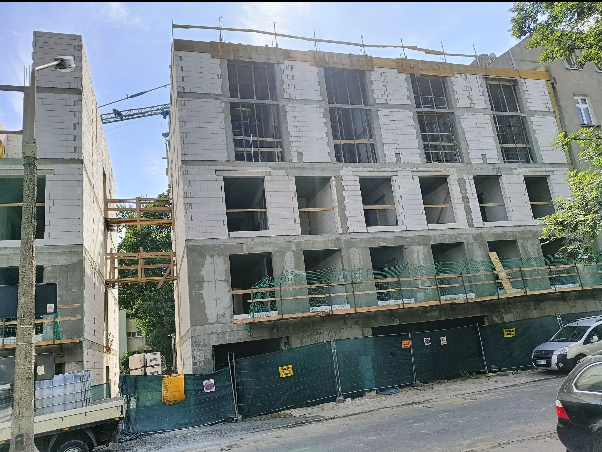 Budowa bloku z białych pustaków, widok z ulicy, z zabezpieczeniami. Częściowo widoczny dźwig i zaparkowane auta. Etap murowania ścian.