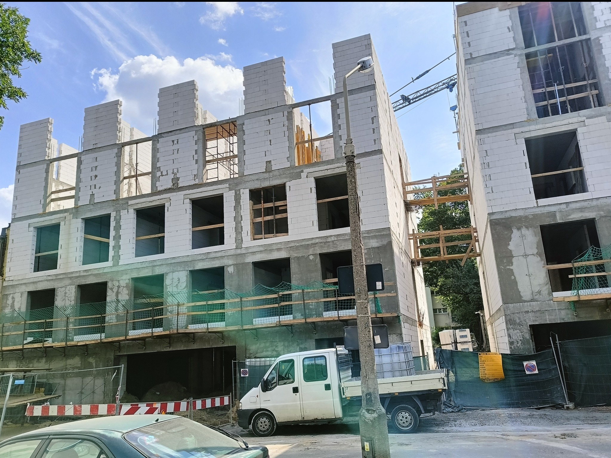 Budowa trzypiętrowego budynku z białych bloczków, z widocznymi otworami okiennymi i elementami rusztowań, na tle błękitnego nieba z chmurami.