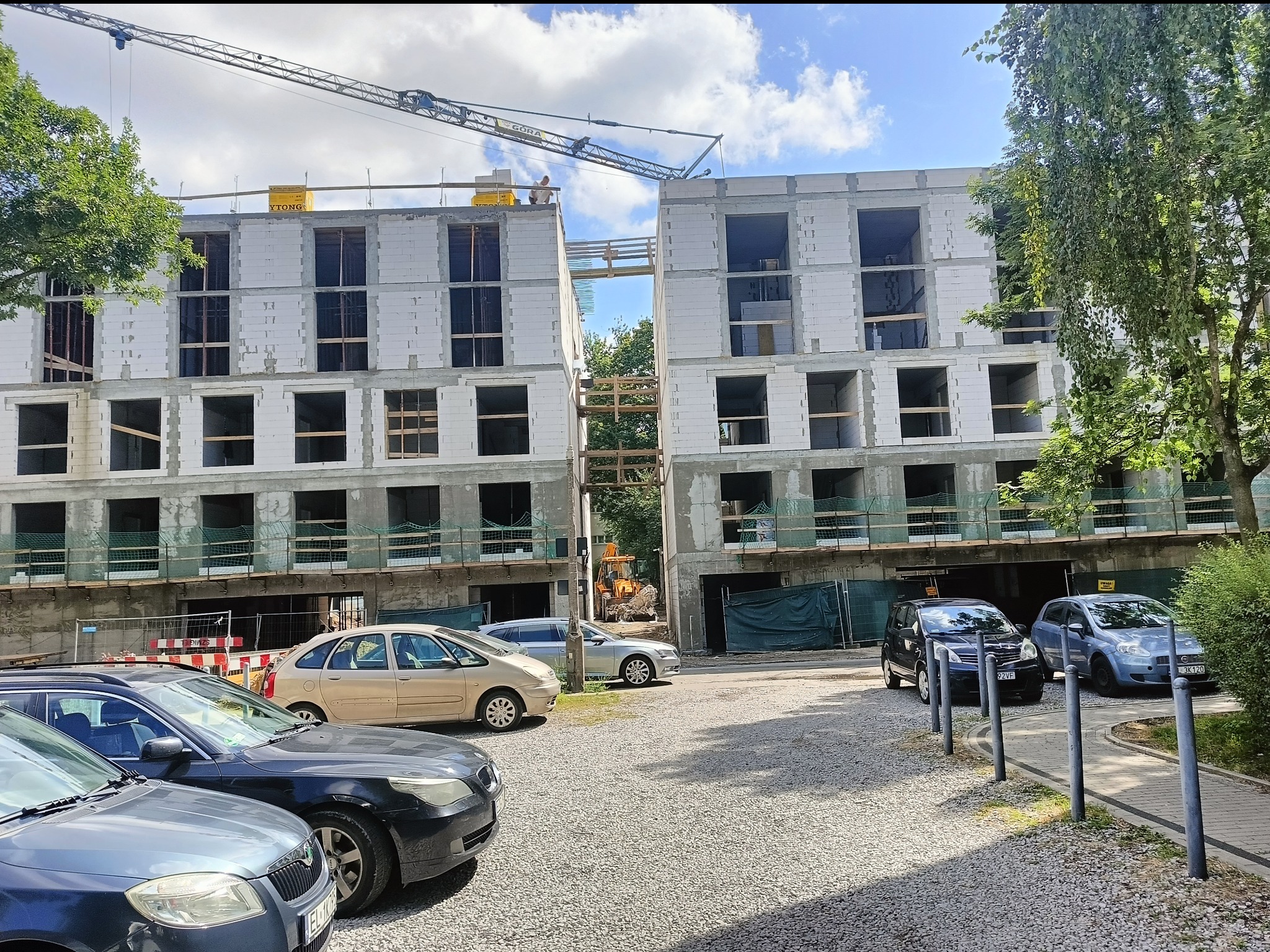 Budowa dwusegmentowego budynku z betonu komórkowego. Widoczny dźwig budowlany, zaparkowane samochody i elementy infrastruktury. Jasne niebo z chmurami w tle.