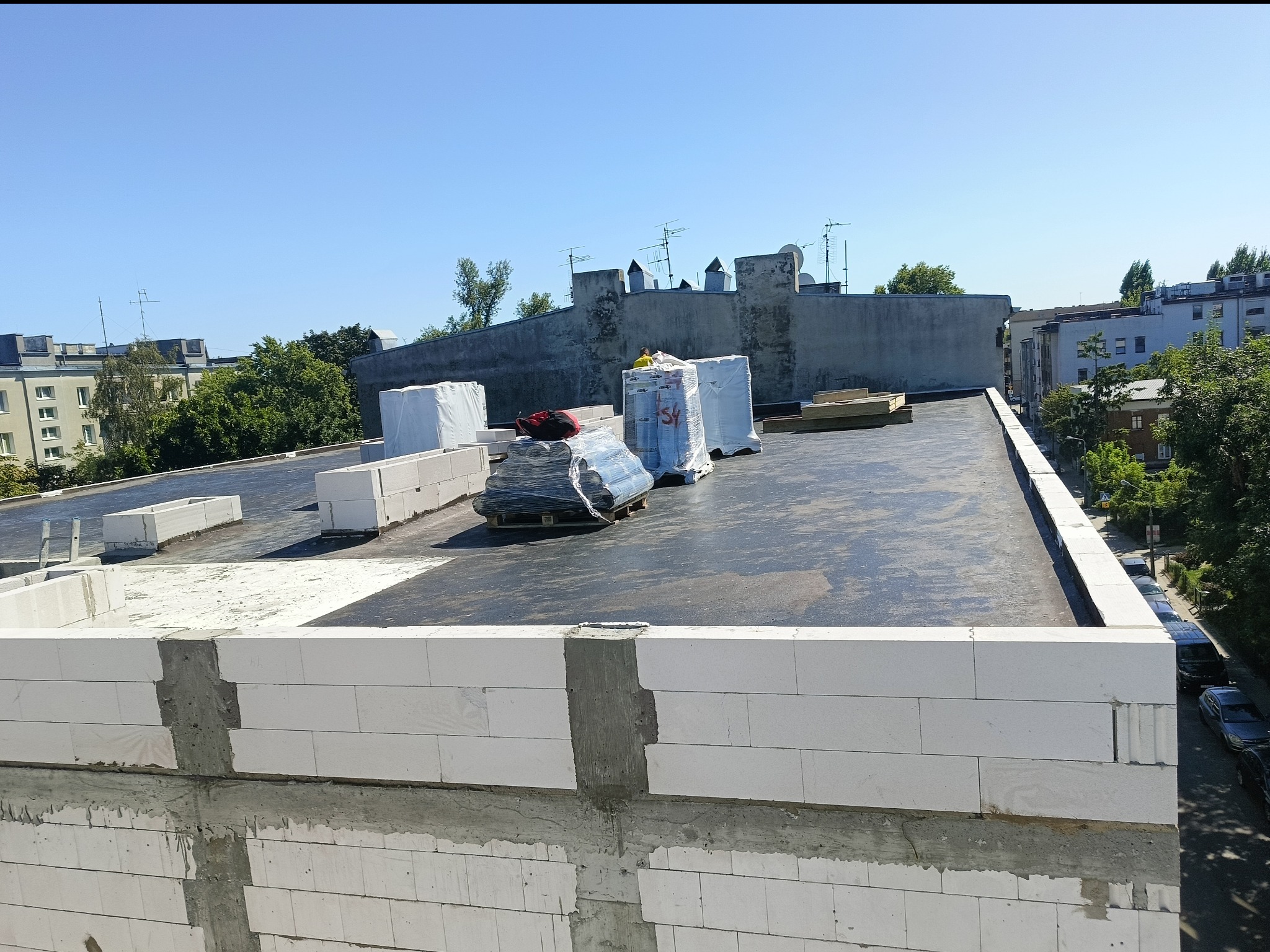 Wznoszenie muru z białych bloczków na dachu budynku. Widoczne materiały budowlane i panorama miasta w tle. Świeże spoiny cementowe.