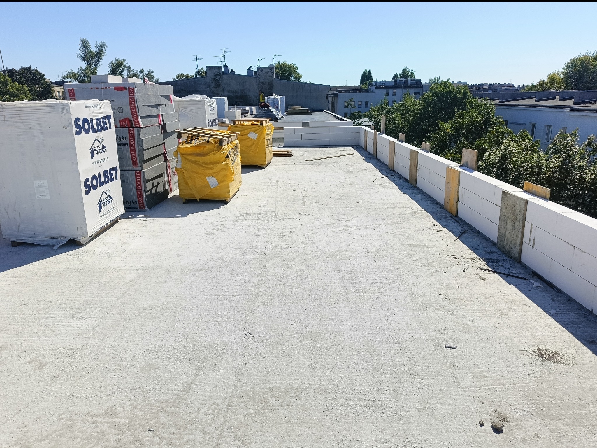 Wznoszenie muru z białych bloczków Solbet na betonowej powierzchni. Palety z materiałami budowlanymi w tle, błękitne niebo nad budową.