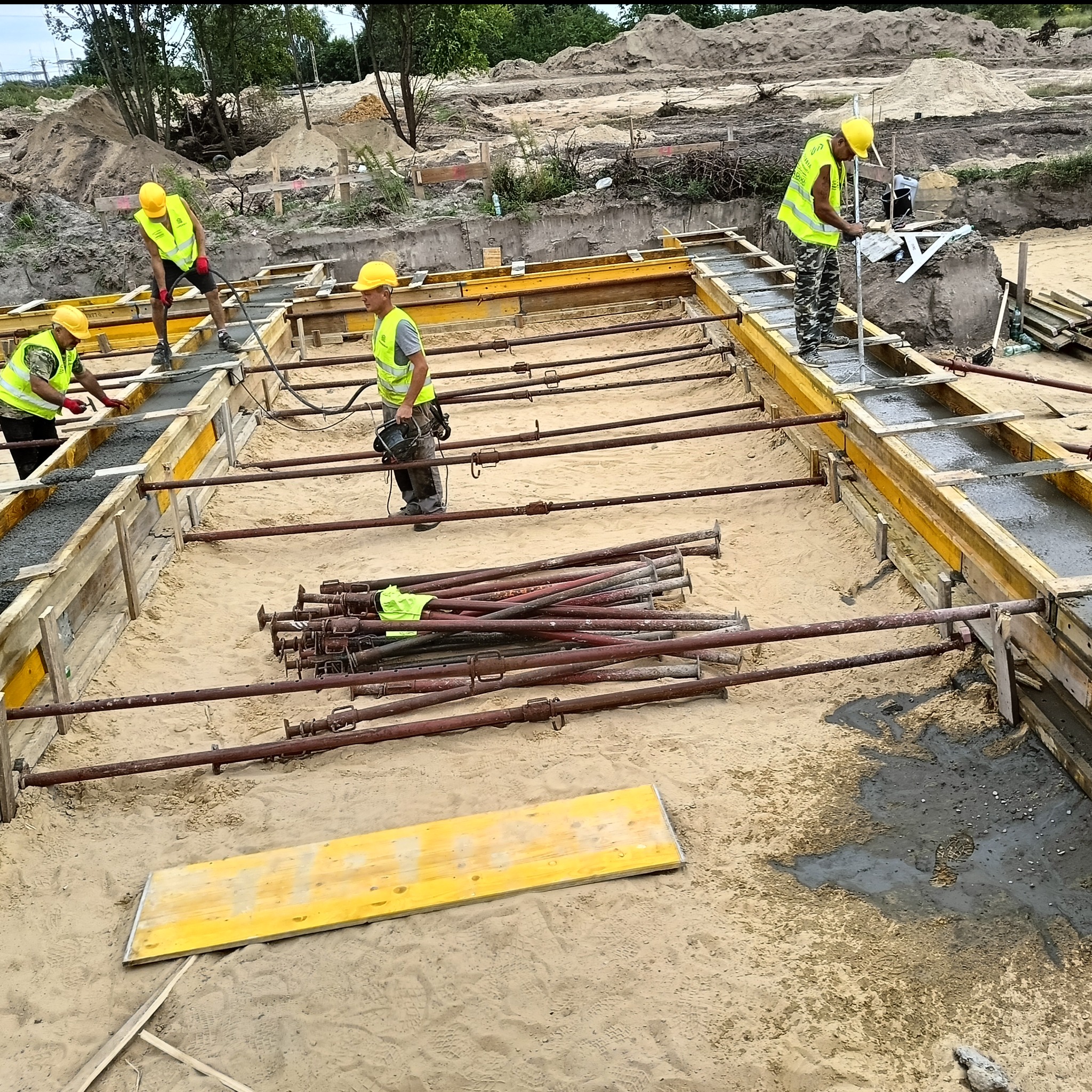 Wylewanie fundamentów: czterech pracowników w żółtych kaskach i kamizelkach wylewa beton w szalunek. Na piasku leżą podpory i deski.