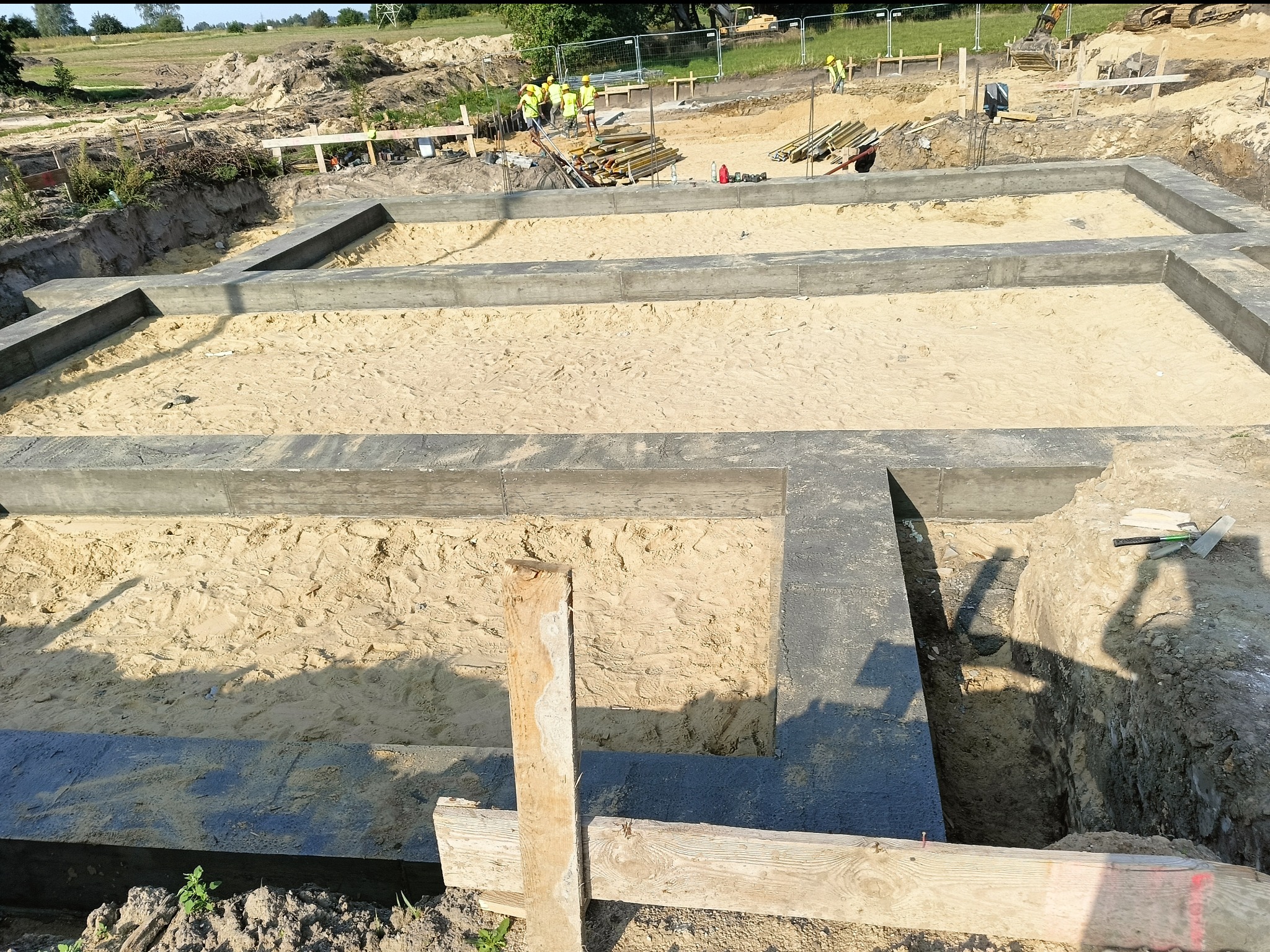 Zalane betonem fundamenty domu, widok z góry na piasek wewnątrz. W tle ekipa budowlana i sprzęt na placu budowy. Kąt ujęcia podkreśla głębię wykopu.